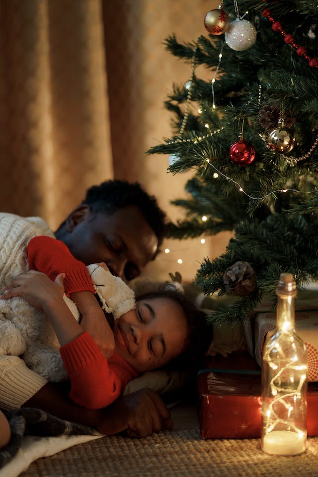 Dad embracing sleeping daughter during holiday season; by a Christmas tree